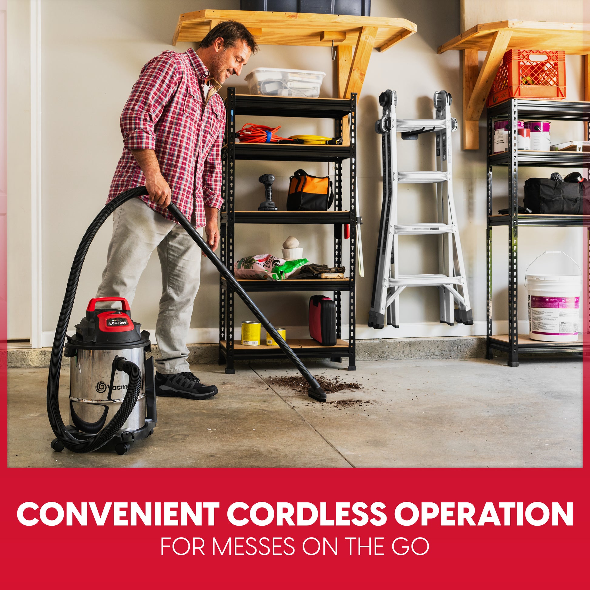 A man in a red plaid shirt and beige pants is using a cordless shop vacuum cleaner in a garage setting. The vacuum has a silver canister body with a red handle and black hose.