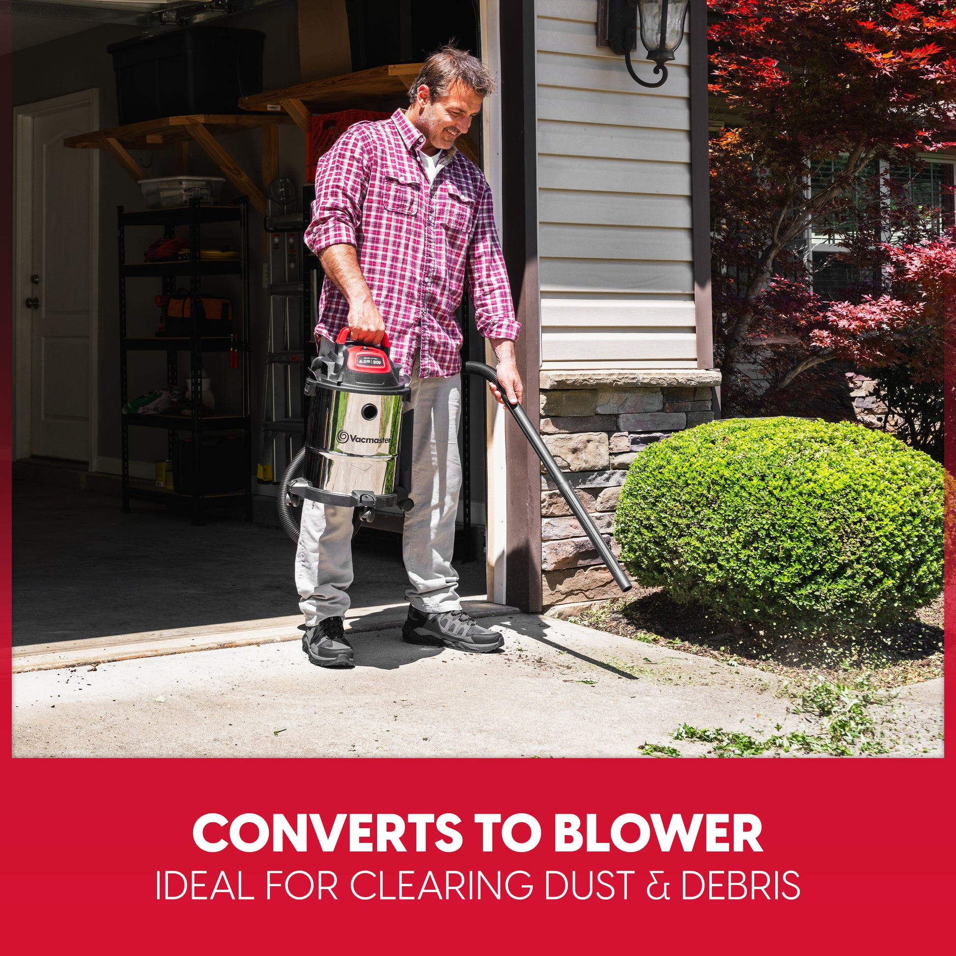 A man in a red plaid shirt and gray pants stands in a garage doorway holding a wet/dry shop vacuum with a long hose attachment, shown clearing dust and debris from a green shrub in a front yard. A red banner at the bottom reads 'CONVERTS TO BLOWER IDEAL FOR CLEARING DUST & DEBRIS'.
