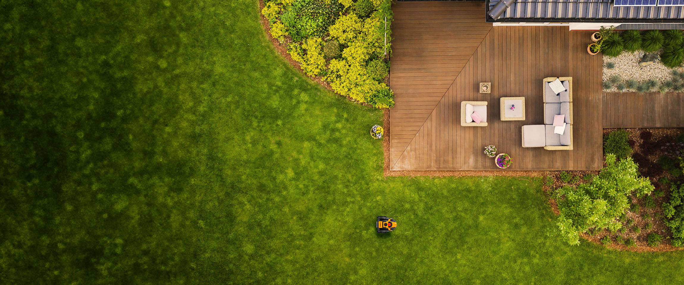 Aerial view of a backyard with a LawnMaster Ocumow robot mower, a wooden deck, outdoor furniture, and green grass.