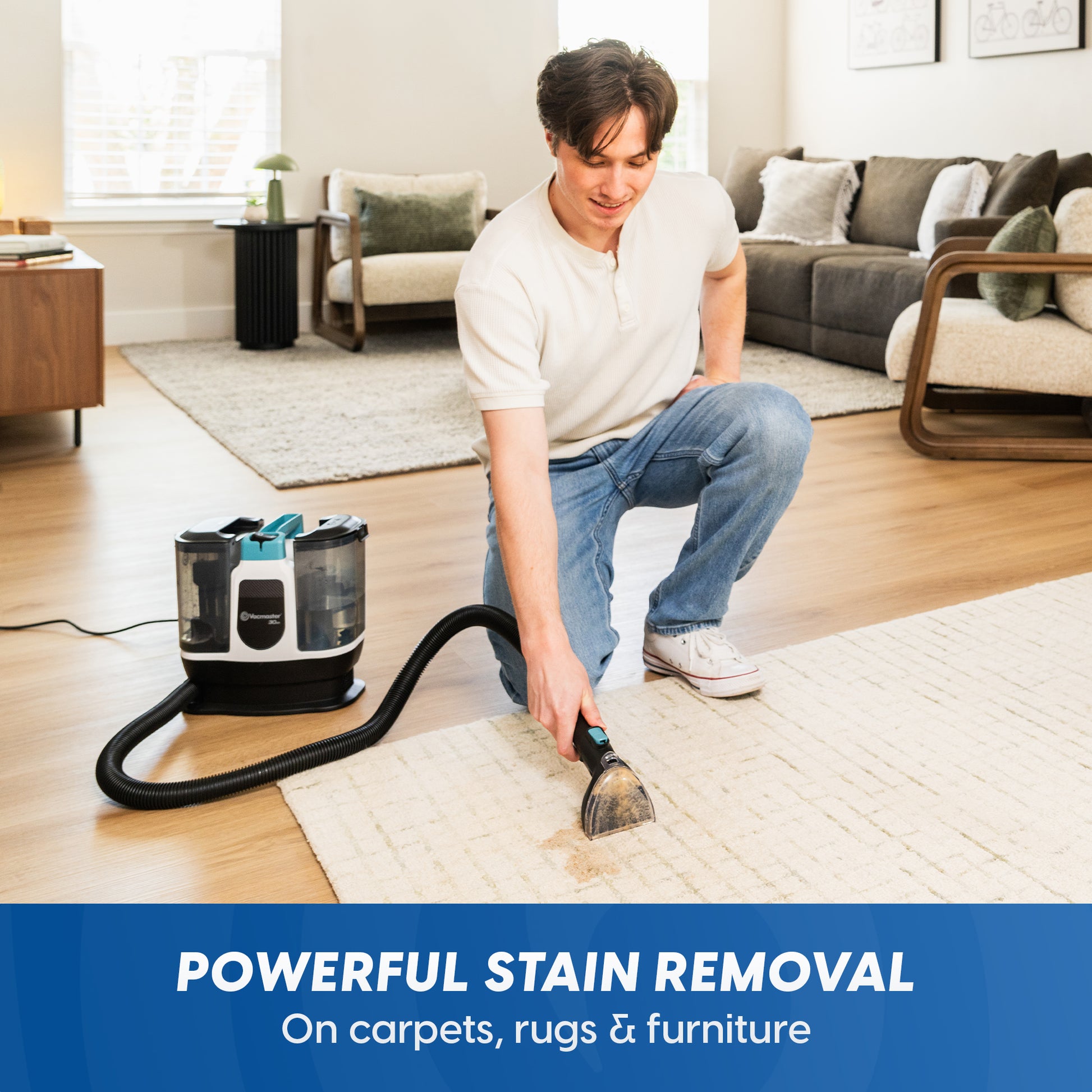 A man kneels on a light beige rug in a bright living room, using a cordless carpet cleaner with a black hose and brush head to clean the rug. The cleaner has a black, gray, and teal body.