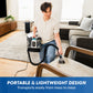 A man kneeling in a living room uses a Vacmaster® 30 Series Mini Carpet & Upholstery Cleaner on a light-colored sofa cushion. The cleaner has a black and white body with two transparent dust bins.