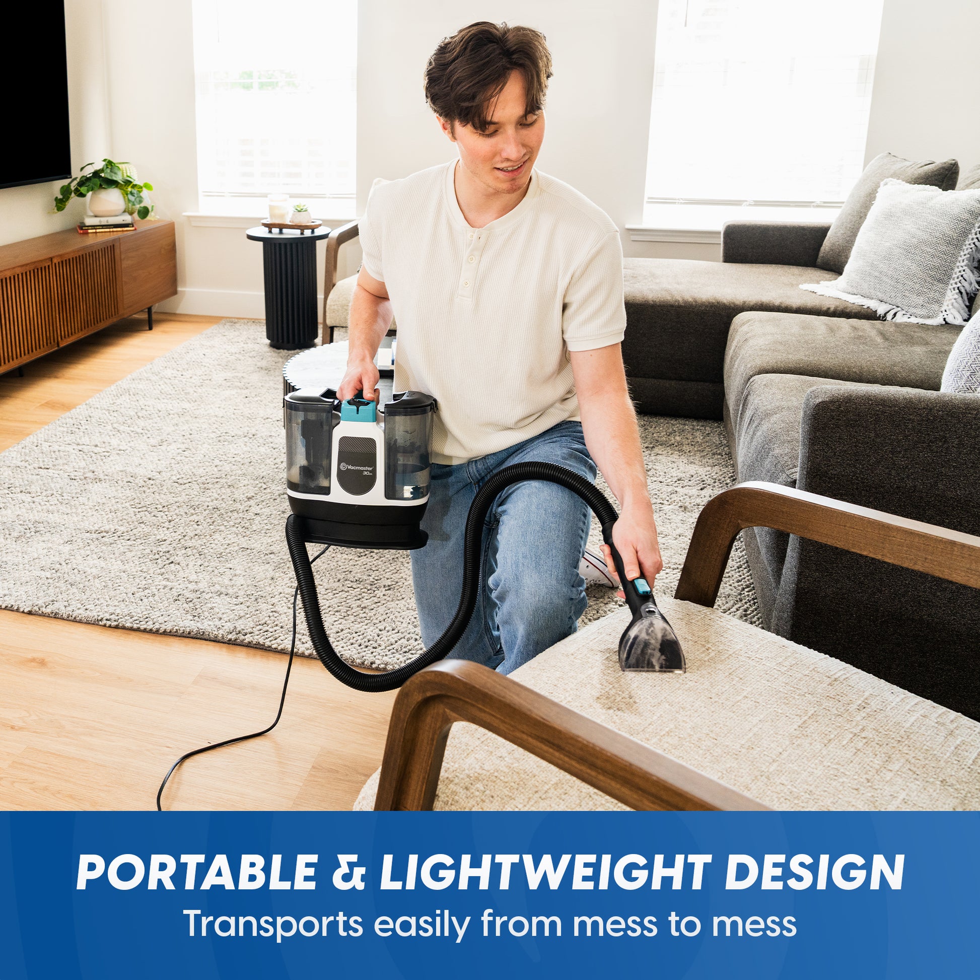 A man kneeling in a living room uses a Vacmaster® 30 Series Mini Carpet & Upholstery Cleaner on a light-colored sofa cushion. The cleaner has a black and white body with two transparent dust bins.