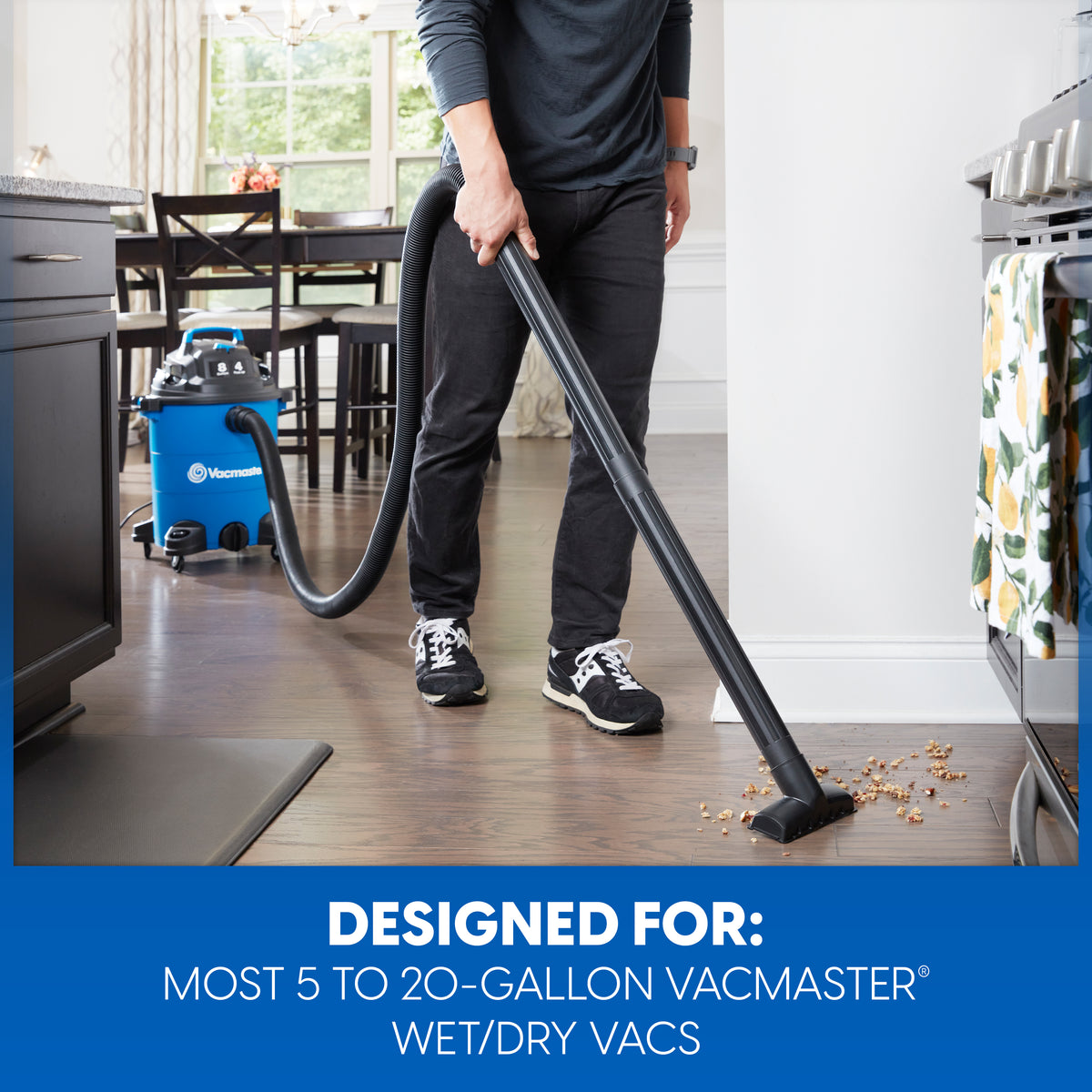 A person in a kitchen using a black vacuum wand attachment connected to a blue and black Vacmaster wet/dry vacuum. A blue text banner at the bottom reads 'DESIGNED FOR: MOST 5 TO 20-GALLON VACMASTER WET/DRY VACS'.