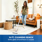 A woman in a living room using a cordless stick vacuum cleaner on a patterned area rug. The vacuum has a white body with blue and silver accents.