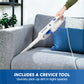 A handheld cordless vacuum cleaner being used on a gray fabric sofa, with a person's hand and arm visible holding it. The vacuum is white with blue accents, and the scene includes a blue throw pillow and a text banner at the bottom that reads 'INCLUDES A CREVICE TOOL Quickly pick up dirt in tight spaces.