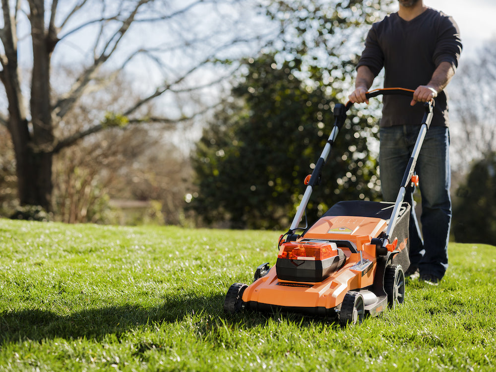 Person using a LawnMaster orange lawn mower on a grassy area with trees in the background